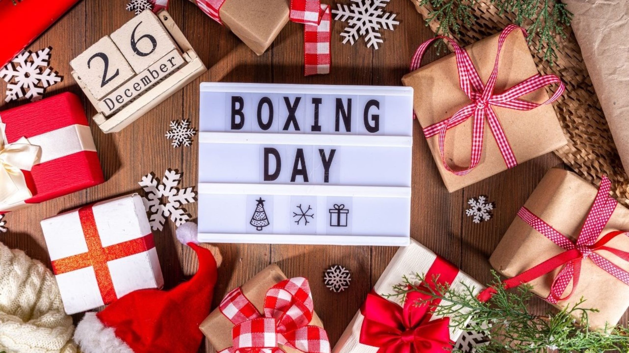 Boxing Day flat lay with wrapped Christmas gifts, festive decorations, snowflakes, and lightbox sign reading Boxing Day on wooden background.