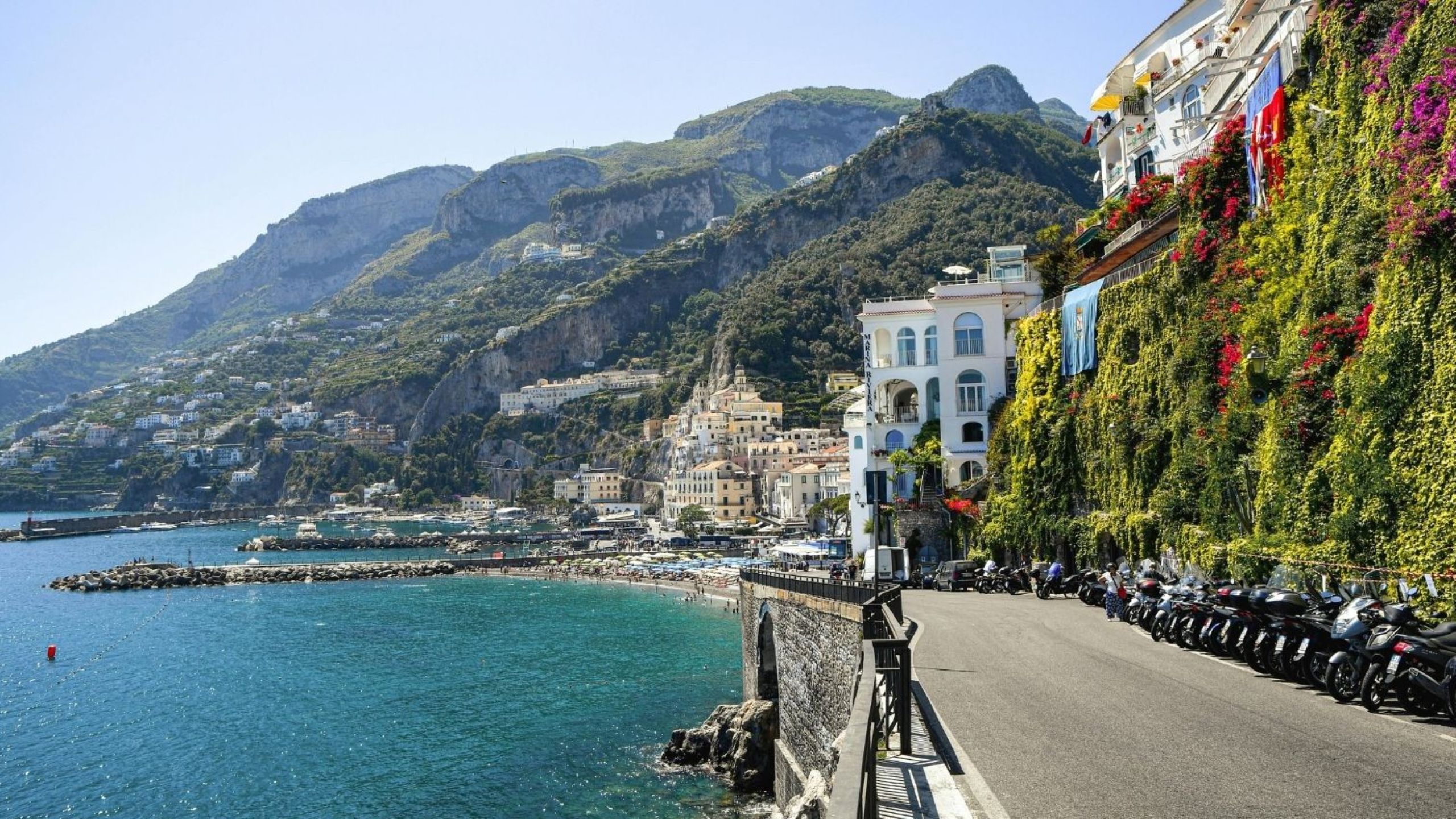Coastal cliffside town with white buildings, greenery, and a row of scooters along a waterfront road by the blue sea at sea level.