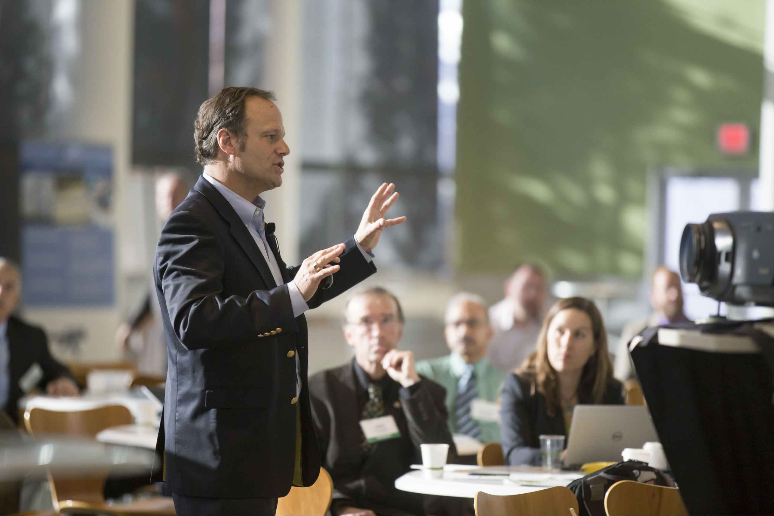 Male speaker in a dark suit gesturing while presenting to a seated audience in a conference room.