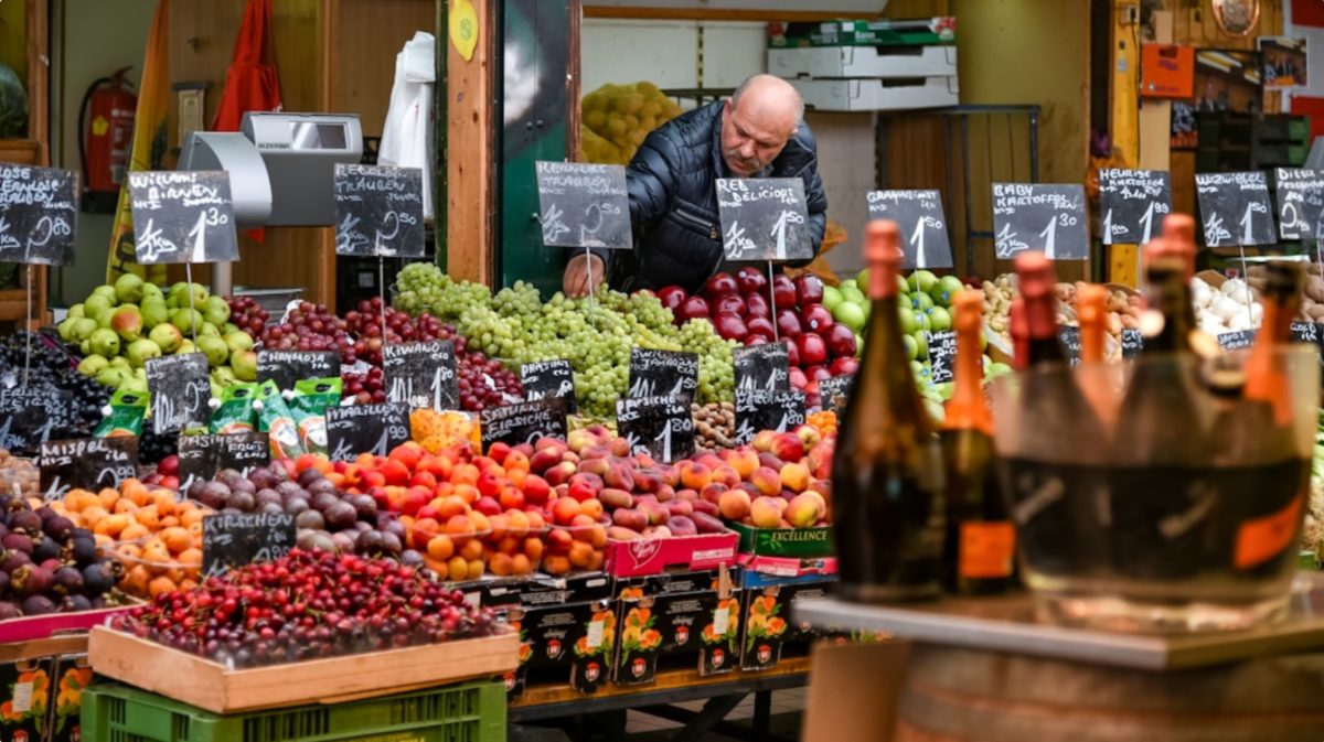 A woman selecting fresh seasonal vegetables at an EU summer farmers market in a sunlit town square.